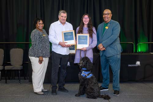Standing on a stage: Ebony Green, Ross M. Chowles, Jenna Baker, Kwesi Brookins and Henry (facility dog)